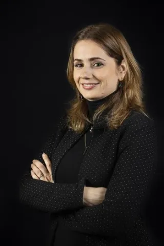 Portrait photographique d’une personne cadrée en buste, portant une veste sombre avec les bras croisés, sur un fond uni noir.