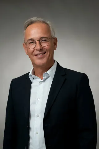 Portrait photographique d’une personne cadrée en buste, portant une veste sombre sur une chemise claire et des lunettes, sur un fond dégradé clair.
