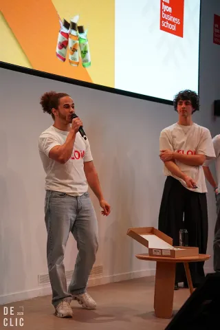 Deux étudiants sur une scène présentent un projet devant un écran de projection, l’un tenant un micro, avec une table et des documents à proximité. 