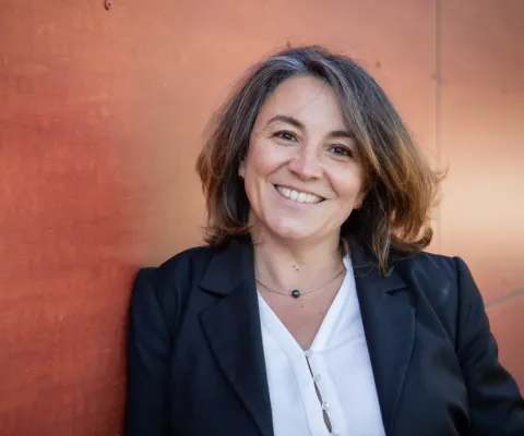 Portrait photographique d’une personne cadrée en buste, portant une veste sombre sur un haut clair, se tenant devant un mur de couleur rouge-orangé.