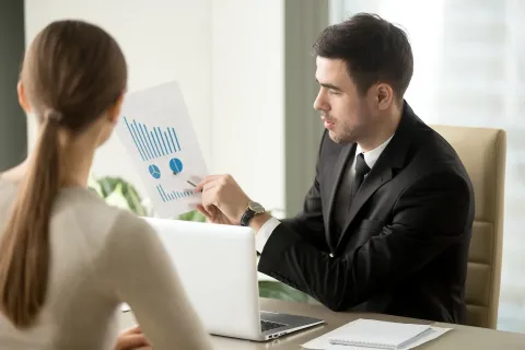 Two people seated at a desk review a document with charts together, with an open laptop in front of them.