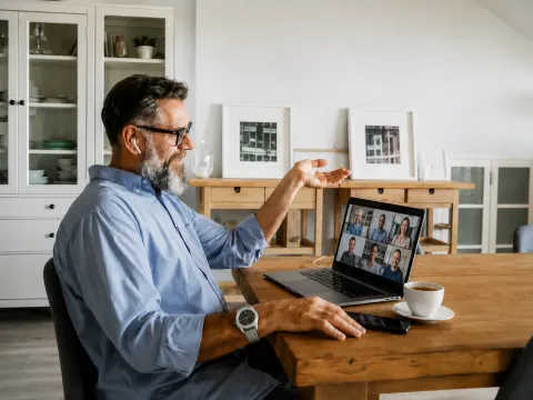 Une personne est assise à une table en bois dans un intérieur, utilisant un ordinateur portable affichant une visioconférence avec plusieurs participants, avec une tasse et un téléphone posés à proximité