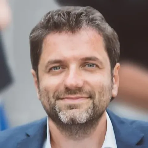 Portrait photographique d’une personne cadrée en buste, portant une veste sombre, sur un fond extérieur légèrement flouté.