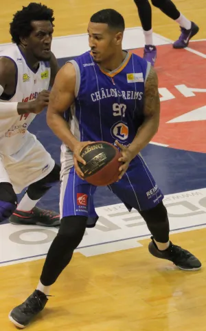 Deux joueurs sur un terrain de basketball, l’un tenant un ballon et pivotant tandis que l’autre se positionne en défense, avec des marquages de terrain visibles au sol.