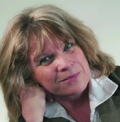 Portrait photographique d’une personne cadrée en buste, avec les cheveux mi-longs, portant une veste sombre sur une chemise claire, la tête appuyée sur une main, sur un fond uni clair.