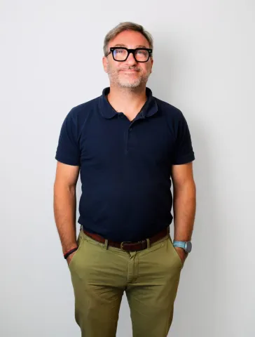 ne personne debout, cadrée en pied, portant un polo foncé et un pantalon clair, avec les mains dans les poches, devant un fond uni gris.