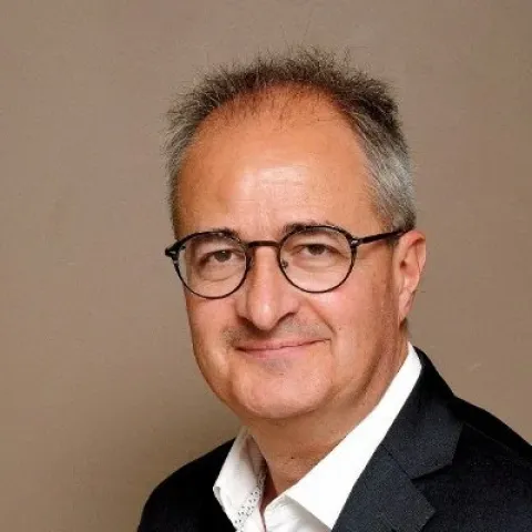 Portrait photographique d’une personne cadrée en buste, portant des lunettes et une veste sombre sur une chemise claire, sur un fond uni beige.