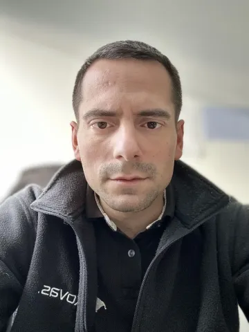 Portrait photographique d’une personne cadrée en buste, portant une veste sombre avec fermeture zippée, dans un intérieur avec un fond clair légèrement flouté.