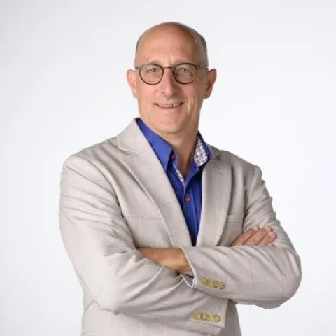 Portrait photographique d’une personne portant des lunettes, une veste claire sur une chemise bleue, les bras croisés, sur un fond uni clair.