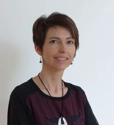 Portrait d’une personne aux cheveux courts portant un haut foncé et un collier, devant un fond clair uni.