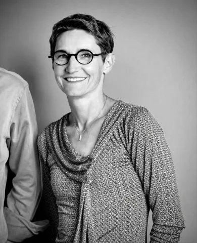 Portrait photographique en noir et blanc d’une personne portant des lunettes, cadrée jusqu’à la taille, vêtue d’un haut à motif et d’un collier, sur un fond uni clair.