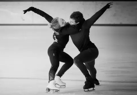 Deux personnes en patins à glace exécutant une figure sur une patinoire intérieure, bras étendus et corps inclinés.
