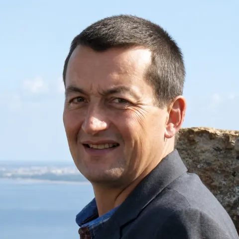 Portrait photographique d’une personne cadrée en buste, portant une veste sombre, en extérieur, avec un plan d’eau et un mur en pierre en arrière-plan.