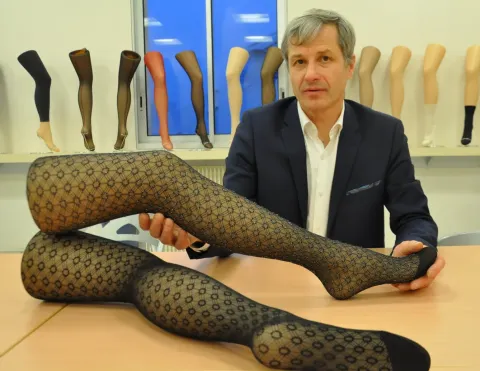 Une personne est assise à une table et tient un objet en forme de jambe recouvert d’un textile à motif, avec plusieurs modèles de jambes présentés sur une étagère en arrière-plan.