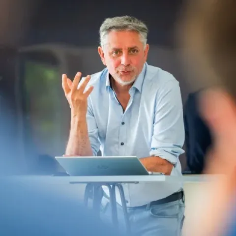 Portrait d’une personne portant une chemise noire à manches longues, debout devant un mur bleu à panneaux décoratifs.
