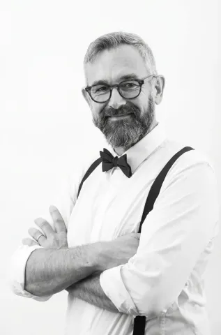 Portrait photographique en noir et blanc d’une personne portant des lunettes, un nœud papillon et des bretelles, les bras croisés, sur un fond uni clair.