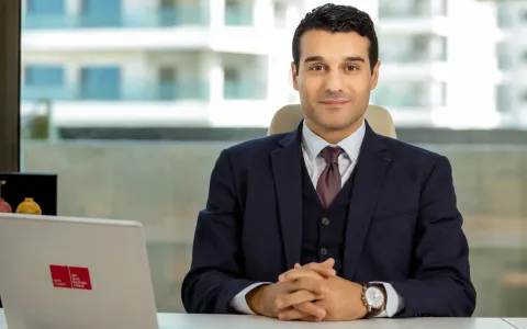 Une personne est assise à un bureau, les mains jointes devant elle, portant un costume sombre et une cravate, avec un ordinateur portable visible au premier plan et de grandes fenêtres en arrière-plan.