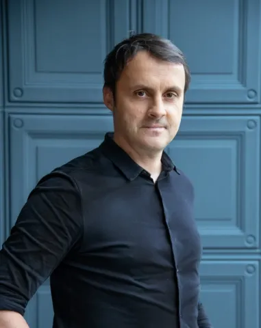 Portrait photographique d’une personne cadrée jusqu’à la taille, portant une chemise sombre, devant un panneau bleu composé de motifs rectangulaires.