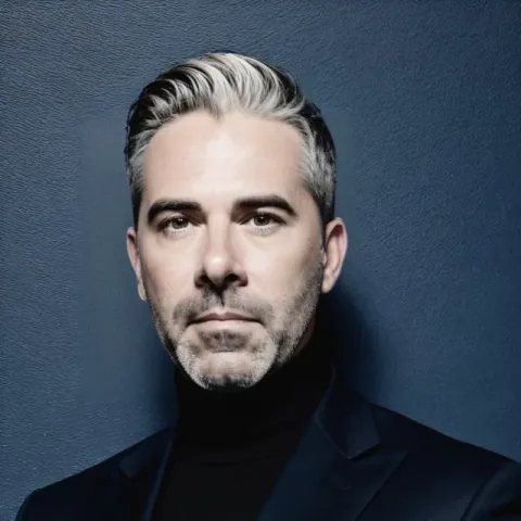 Portrait d’une personne portant un col roulé noir et une veste sombre, devant un fond bleu foncé uni.