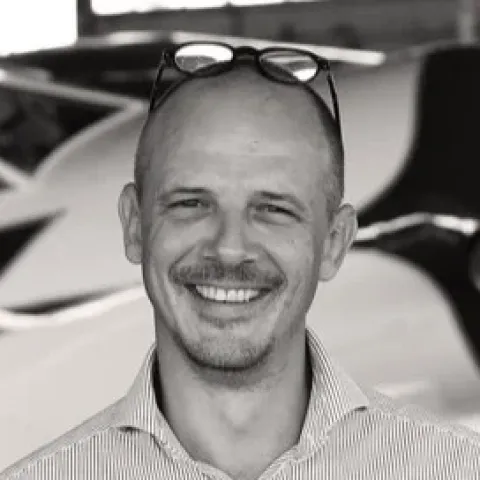 Portrait photographique en noir et blanc d’une personne cadrée en buste, portant une chemise à fines rayures, avec une paire de lunettes posée sur la tête, sur un fond intérieur flou.