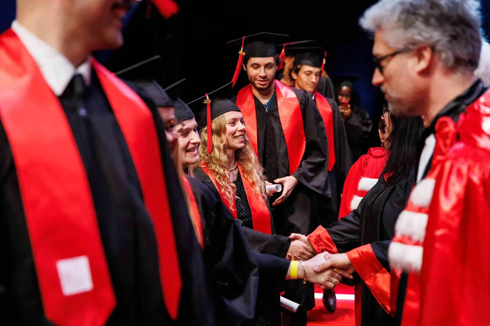 Remise de diplômes : des diplômés en toge noire et écharpe rouge serrent la main d’un représentant sur scène.